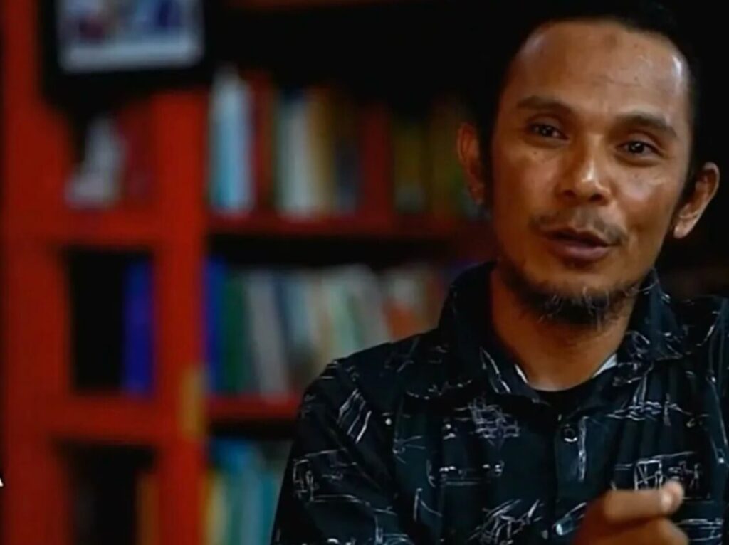 Man with short hair and goatee wearing a dark patterned shirt speaks to camera in a library or bookstore backdrop with shelves behind him